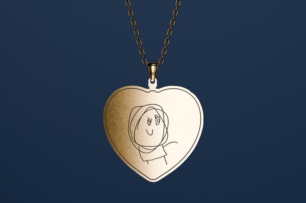 A gold heart-shaped pendant on a fine chain, printed with a child's drawing of a smiling person with big looping hair, hanging against a navy wall