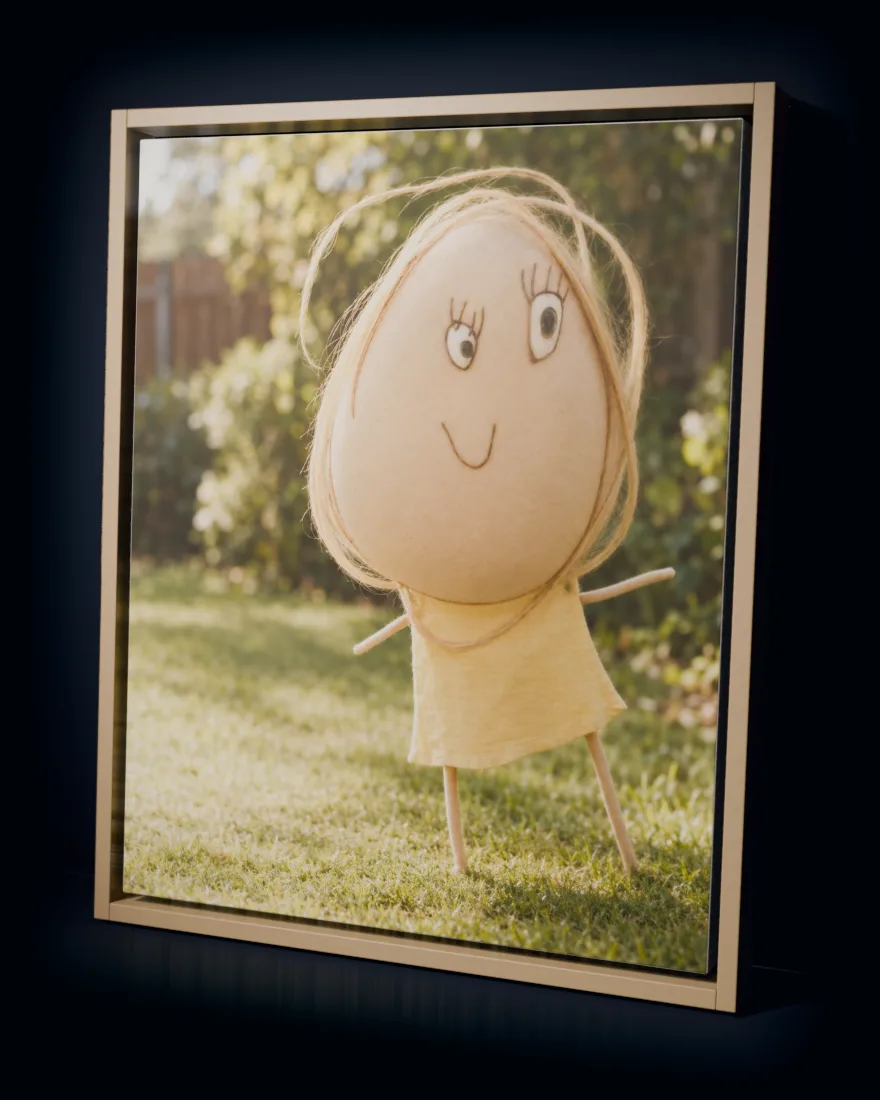 A child’s drawing of a girl with a giant round head, made real with yarn hair and a yellow dress in a sunny garden, printed as a framed canvas in a natural wood frame
