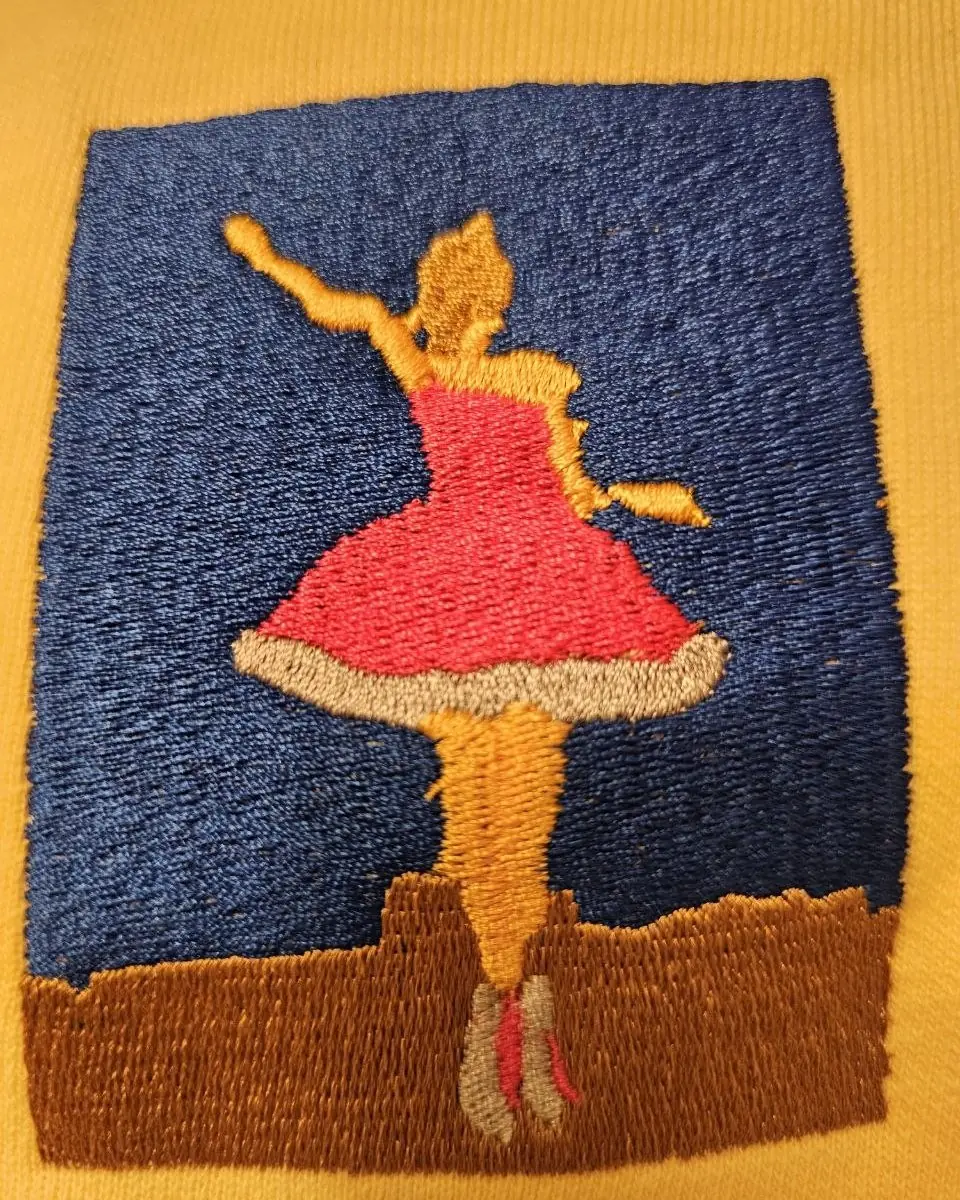 The same ballerina painting embroidered in thread on a butter-yellow sweatshirt, close enough to see the individual stitches