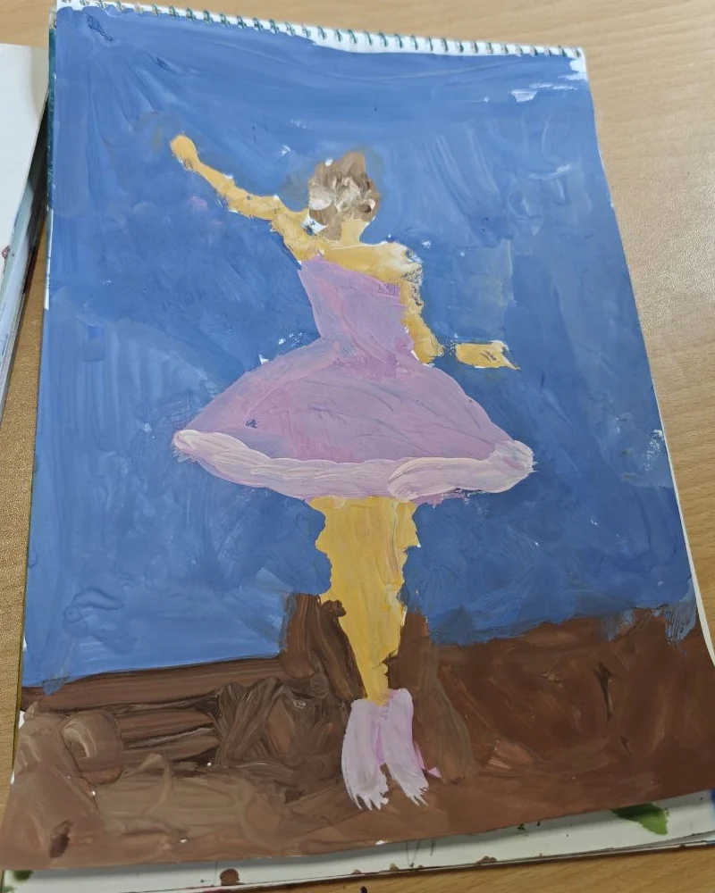 A child's painting of a ballerina in a pink dress against a blue sky, on paper in a spiral sketchbook