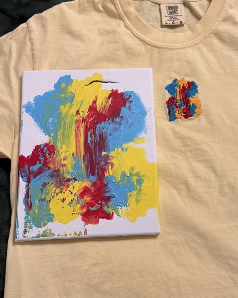A toddler's abstract painting on a small canvas, laid on the butter-yellow t-shirt embroidered with the same painting in thread
