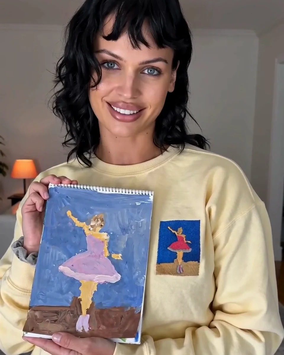 A woman wearing a butter-yellow sweatshirt with a child's ballerina painting embroidered on the chest, holding up the original painting beside it