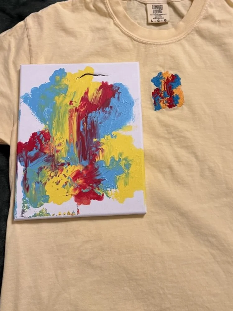 A toddler’s abstract painting on a small canvas, laid on a butter-yellow t-shirt embroidered with the same painting in thread