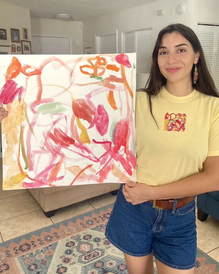A smiling woman holding up a large pink and orange watercolor painting beside her, wearing a butter-yellow t-shirt embroidered with the same painting in thread