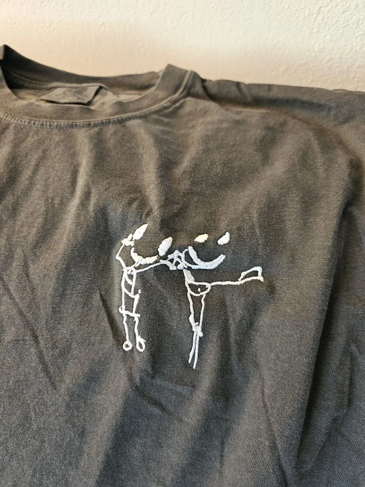 A charcoal-gray t-shirt embroidered with a child’s line drawing of two dancing figures, stitched in white and pale blue thread across the chest